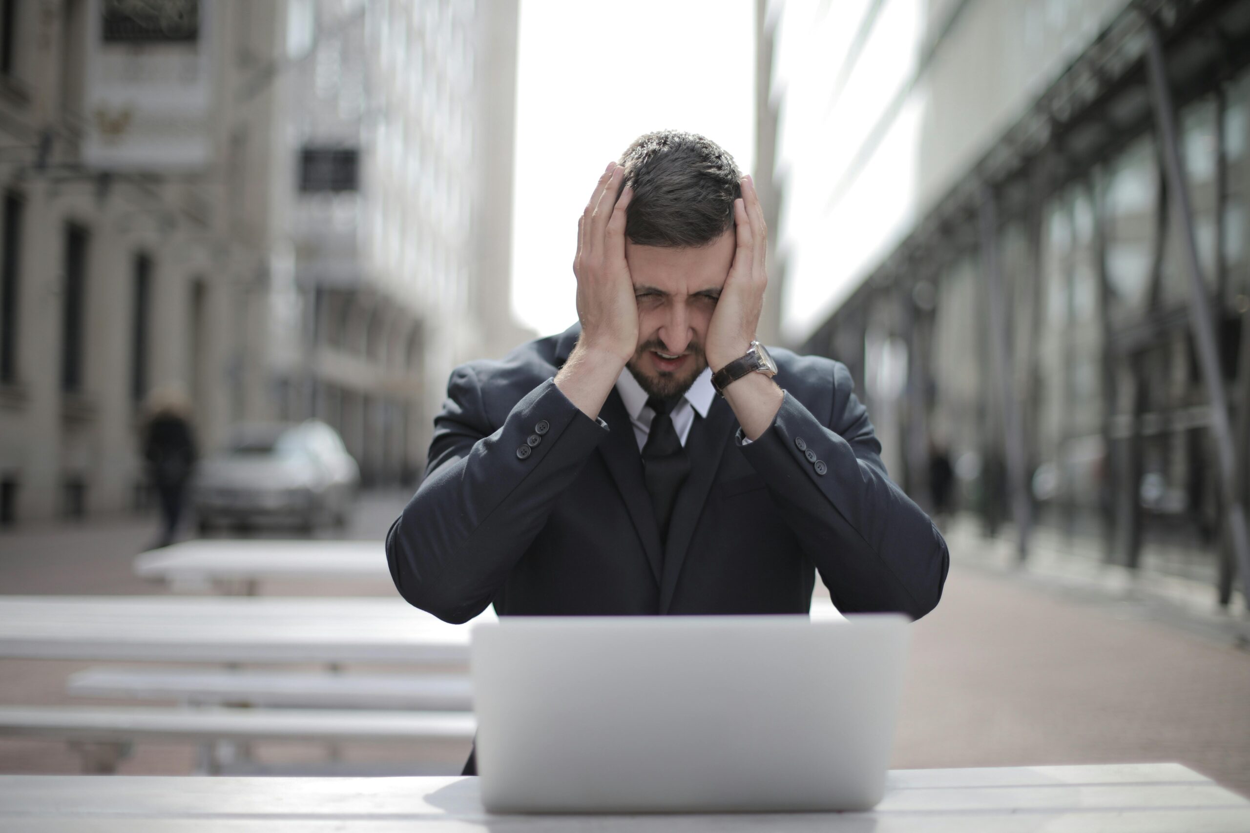 A businessman in a suit looks stressed, sitting outdoors with a laptop in a city environment.