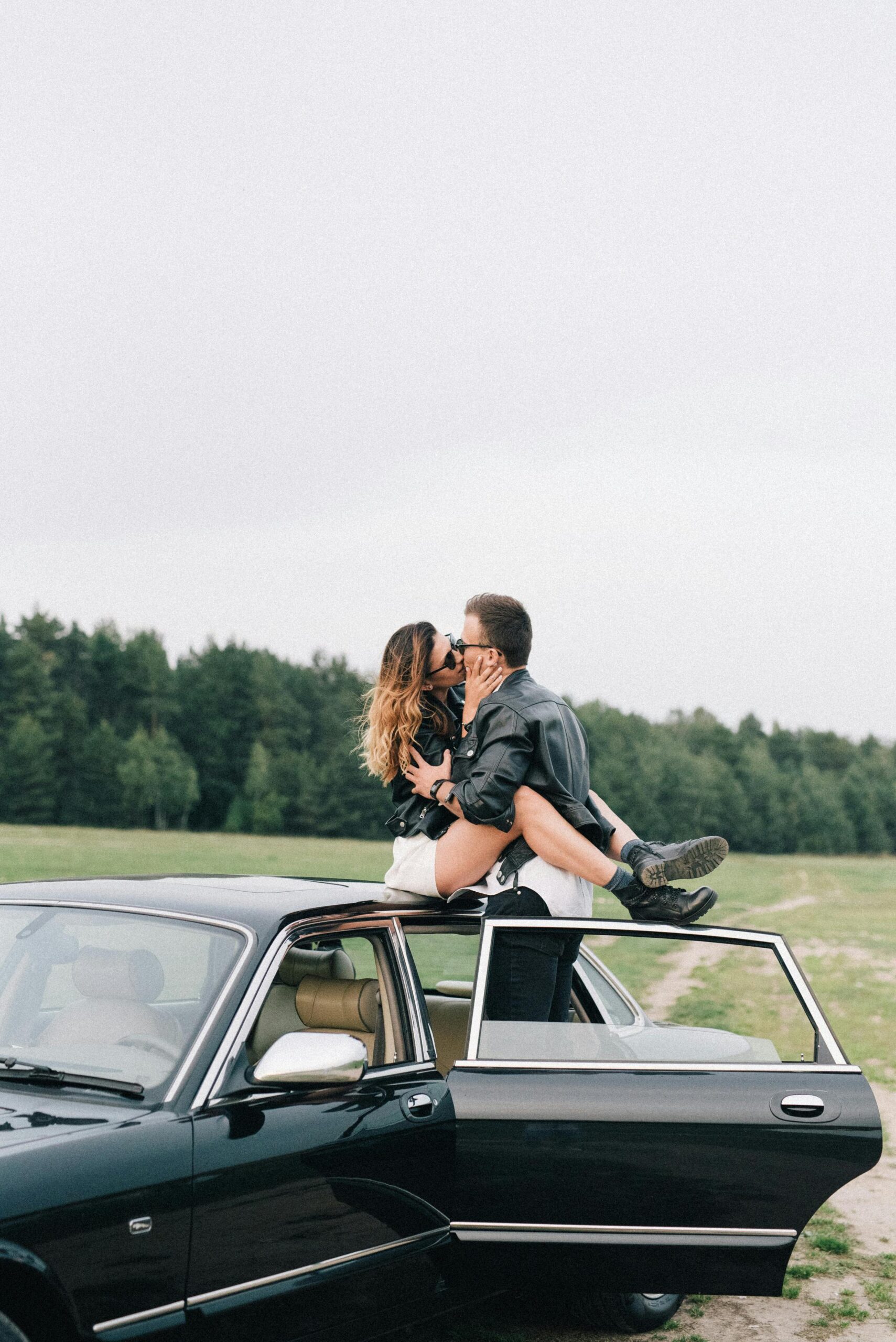 Couple in leather jackets sharing a kiss atop a vintage black car in a scenic outdoor setting.